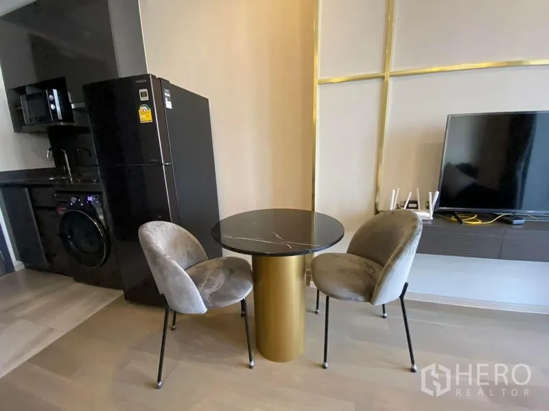 Condominium for rent in Watthana, Bangkok - Compact dining nook with round table, two chairs, wall-mounted TV, and fridge beside the pantry kitchen.