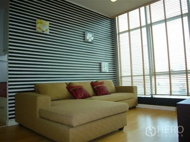 คอนโด ให้เช่าในKhlong San, Bangkok - ห้องนั่งเล่นโซฟา L‑shape ผนังลายขวาง หน้าต่างสูงพร้อมมู่ลี่