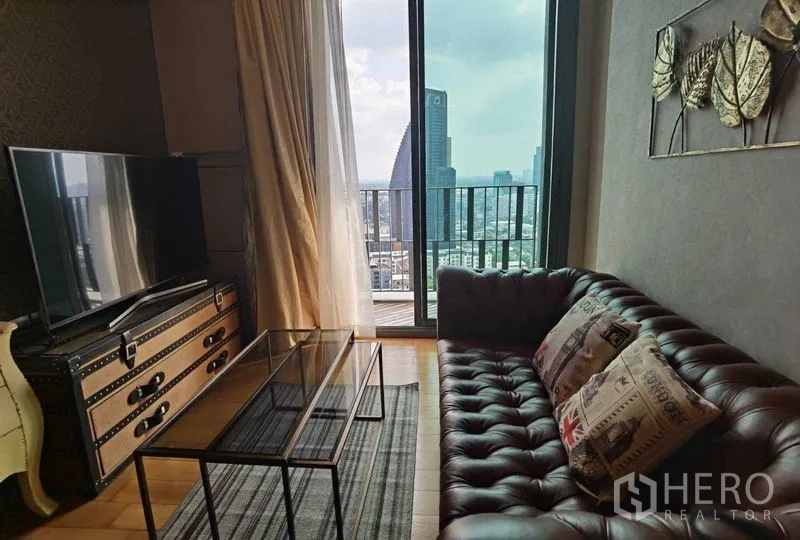 公寓 出租或出售在Khlong Toei, Bangkok - 客厅配皮质沙发、玻璃茶几与电视柜，落地窗连接阳台俯瞰城市。