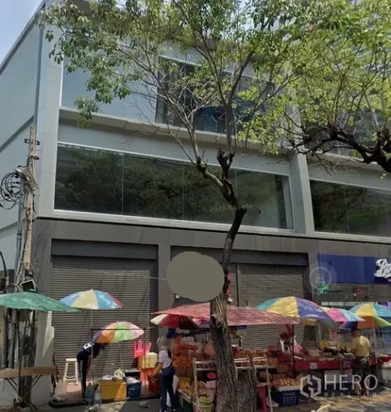 零售空间 出租在Watthana, Bangkok - 临街现代零售楼，大片玻璃窗与卷闸门，路边彩色遮阳伞与行道树，位于曼谷瓦他那区