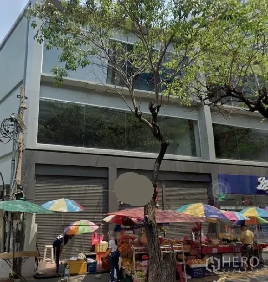 零售空间 出租在Watthana, Bangkok - 临街现代零售楼，大片玻璃窗与卷闸门，路边彩色遮阳伞与行道树，位于曼谷瓦他那区