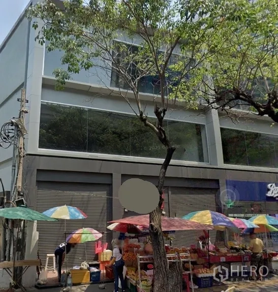 零售空间 出租在Watthana, Bangkok - 临街现代零售楼，大片玻璃窗与卷闸门，路边彩色遮阳伞与行道树，位于曼谷瓦他那区