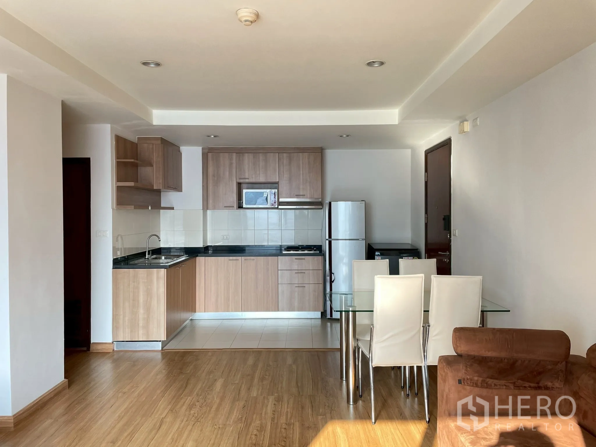 Apartment for rent in Khlong Toei, Bangkok - Open-plan kitchen with wood cabinets, fridge, microwave, and a glass dining table with chairs.