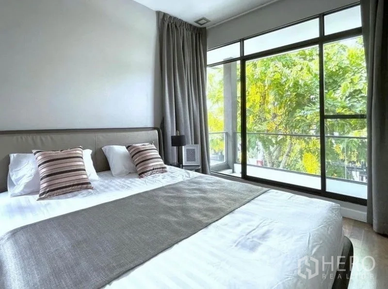 Detached House for rent in Prawet, Bangkok - Master bedroom with king bed, floor‑to‑ceiling windows, grey curtains and leafy outlook.