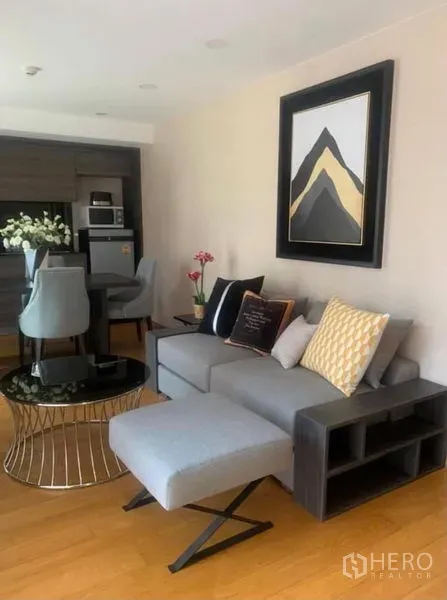Condominium for rent or sale in Pathum Wan, Bangkok - Bright living room with grey sectional sofa, round black coffee table, wall art, and wood floors in a Pathum Wan condo.