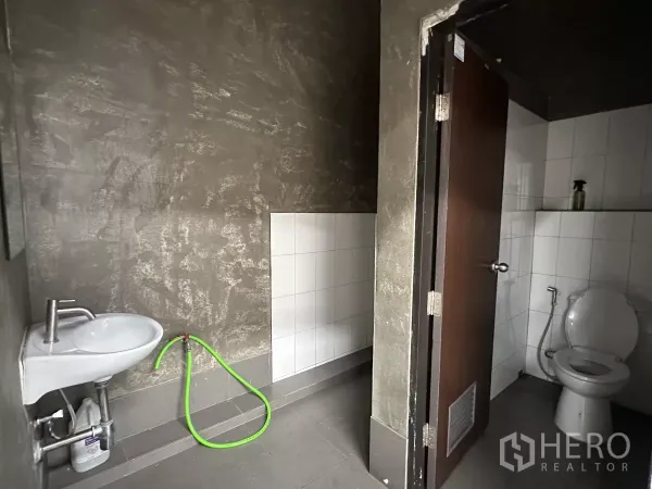 Warehouse for rent in Khlong Toei, Bangkok - Warehouse restroom featuring tiled WC, wall-mounted sink and hose against textured gray walls.
