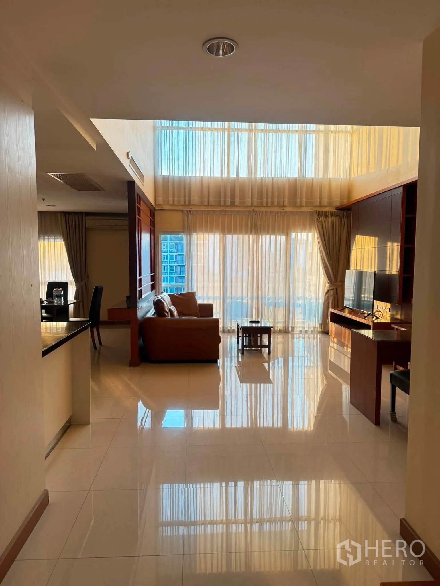 Condominium for rent in Khlong Toei, Bangkok - Open-plan living area with sofa, TV console, glossy tile floors, and double-height windows.