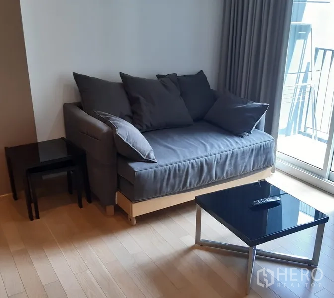 คอนโด ให้เช่าในKhlong Toei, Bangkok - ห้องนั่งเล่นคอนโดสว่าง โซฟาสีเทา โต๊ะกลางกระจก พื้นไม้ และประตูบานเลื่อนออกสู่ระเบียง