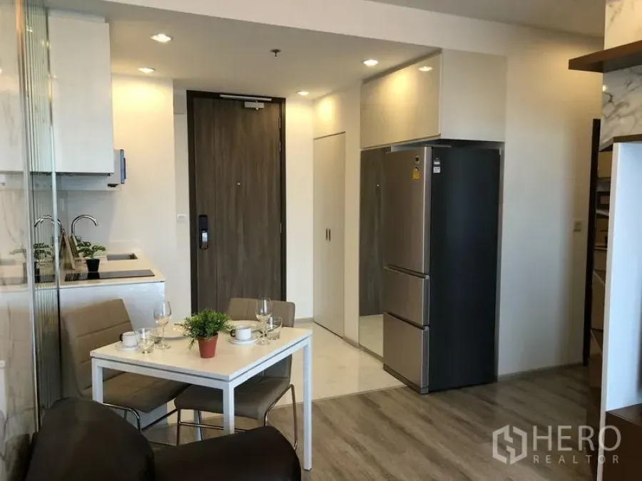 公寓 出租在Bang Na, Bangkok - 开放式厨房与餐区，白色餐桌、嵌入式橱柜及不锈钢冰箱，临近入户门。