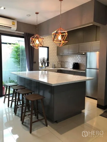 联排别墅 出租在Lat Phrao, Bangkok - 时尚厨房，白色石材中岛、吊灯、高脚凳与灰色柜体，可直达花园。