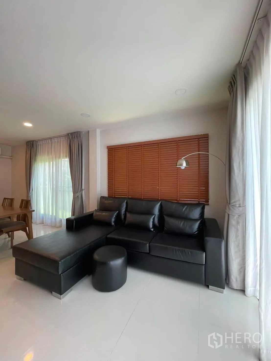 独栋房屋 for rent in Bang Phli, Samut Prakan - 客厅一角放置黑色L形皮沙发与脚凳，落地窗配纱帘与落地灯。