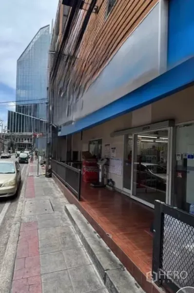零售空间 出租在Watthana, Bangkok - 一层临街商铺外立面，蓝色雨棚与玻璃入口，旁有人行道与近处现代写字楼（Phrom Phong）。