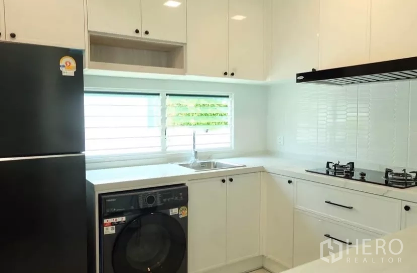 Detached House for rent in Bang Phli, Samut Prakan - Modern white L-shaped kitchen with gas hob, hood, sink, fridge and washing machine under the window.