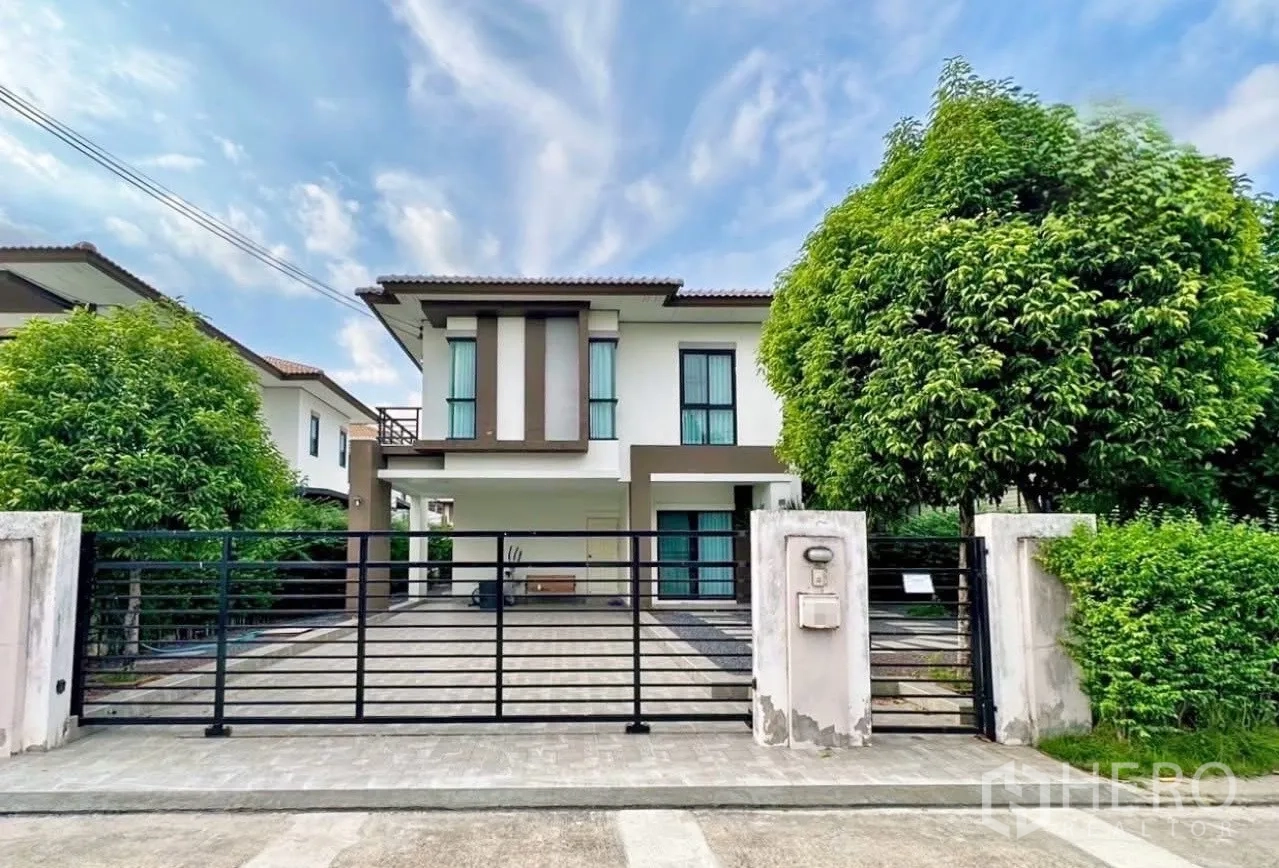 บ้าน ขายในLam Luk Ka, Pathum Thani - บ้านเดี่ยวสองชั้นสไตล์โมเดิร์น พร้อมประตูรั้วเหล็ก ทางเข้ารถกว้างและต้นไม้ใหญ่ท่ามกลางท้องฟ้าแจ่มใส