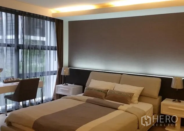Condominium for rent or sale in Watthana, Bangkok - Bright bedroom with large windows, a queen bed, accent wall, bedside tables and a work desk by the window.