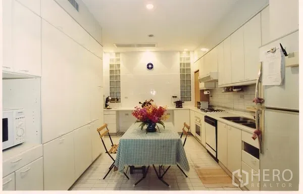 Townhouse for rent or sale in Watthana, Bangkok - Bright galley kitchen with white built-in cabinets, full appliances, and a dining table with flowers.