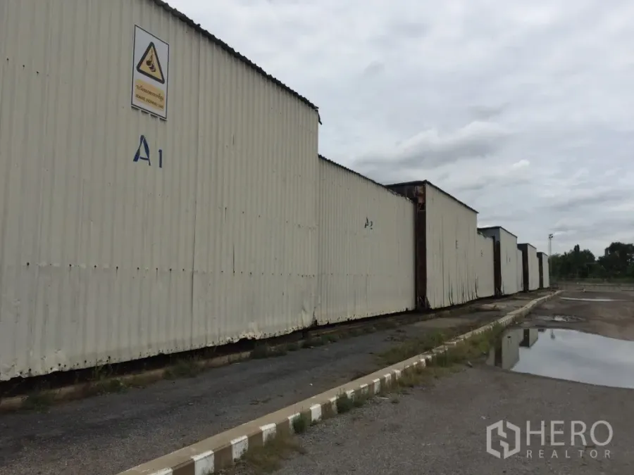 货栈 出租在Mueang Pathum Thani, Pathum Thani - 金属板外墙的仓库排屋，标注A1–A2，旁有宽阔车道与排水沟，阴天景色。