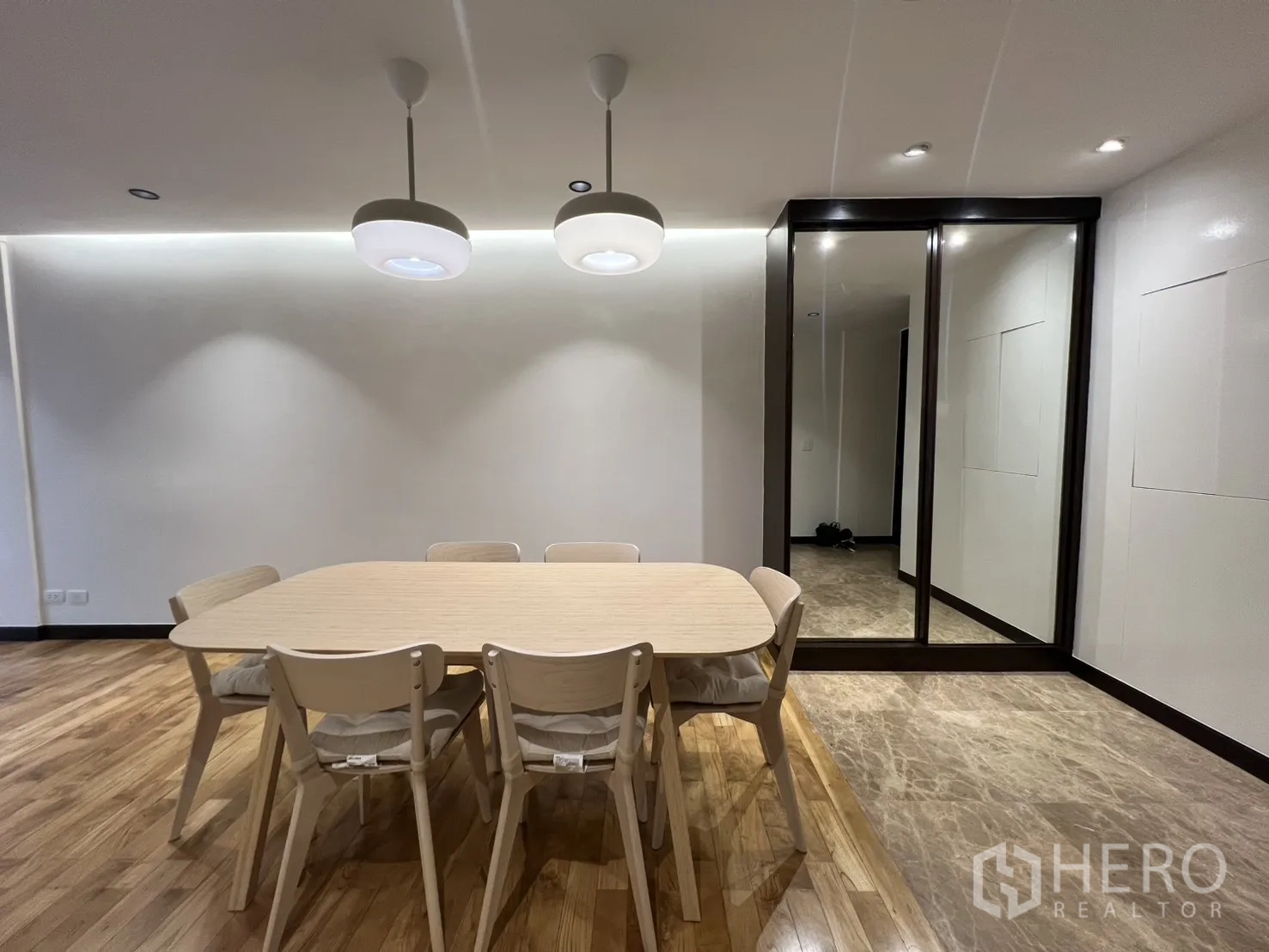 Apartment for rent in Watthana, Bangkok - Six-seat dining table under twin pendant lamps beside a mirrored storage wall and marble-tiled entry.