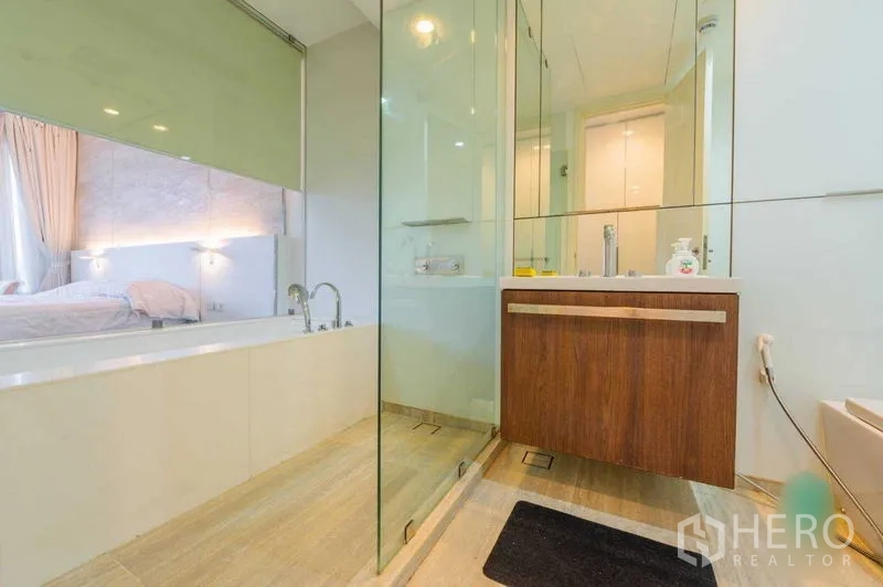 Condominium for rent in Pathum Wan, Bangkok - Bathroom with glass shower, wooden vanity, and a soaking tub adjoining the bedroom behind a sliding glass partition.