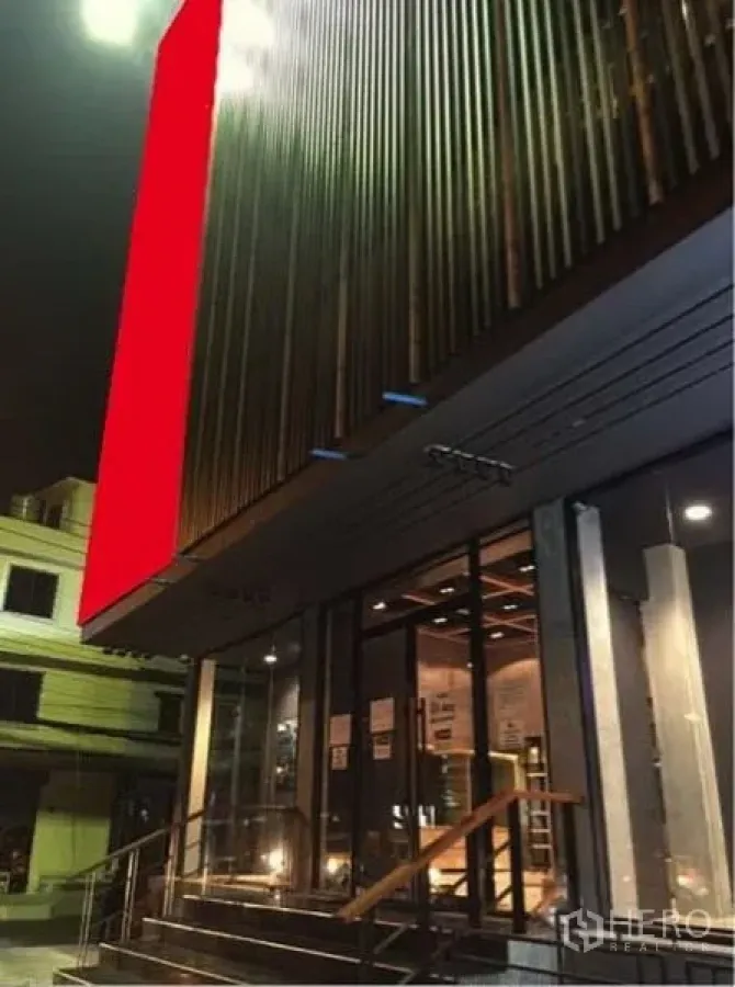 店屋 出租在Suan Luang, Bangkok - 夜景外立面，整面玻璃店面与金属百叶，大幅红色招牌与台阶扶手入口。