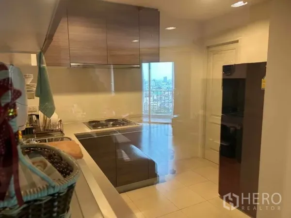 Condominium for sale in Huai Khwang, Bangkok - Separate kitchen with glossy cabinets, electric cooktop, sink and refrigerator, with a city view beyond.