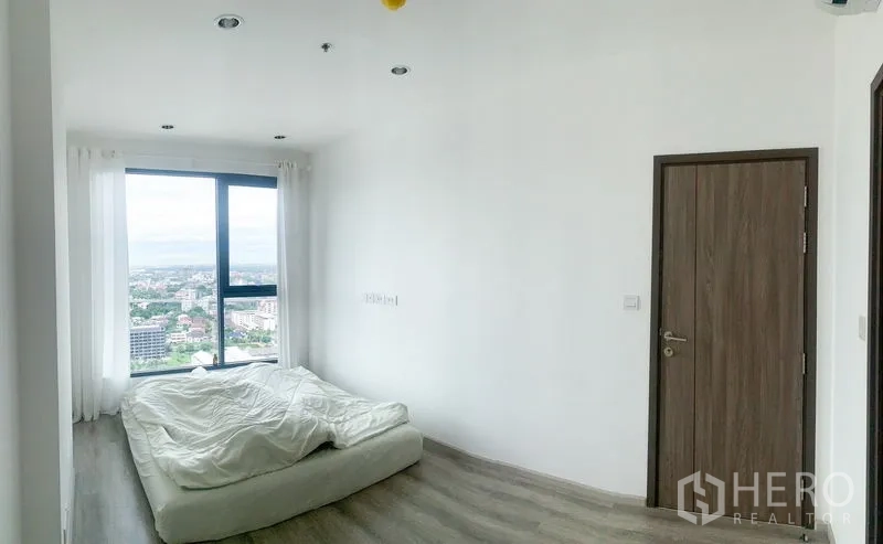 Condominium for sale in Bang Na, Bangkok - Bright bedroom with floor-to-ceiling window, city view, and light wood flooring.