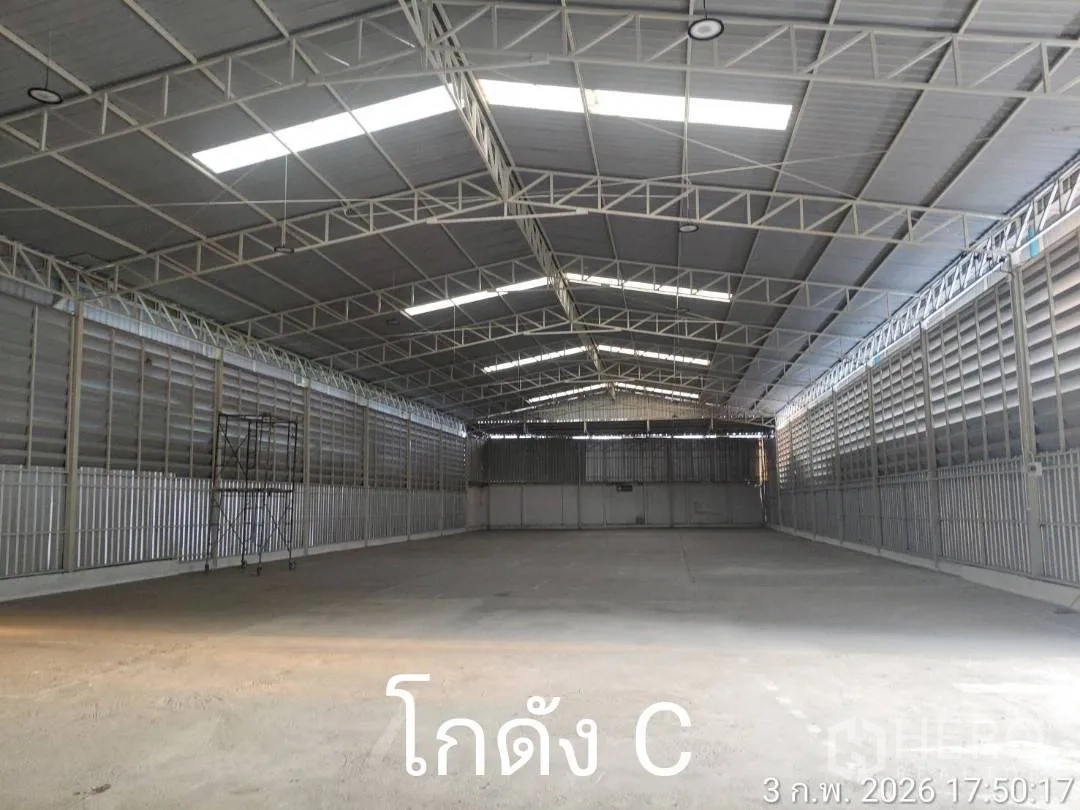 Warehouse for rent in Min Buri, Bangkok - Interior of a warehouse with steel truss roof, skylight panels, ventilated metal walls, concrete floor, and a large roller shutter at the far end.