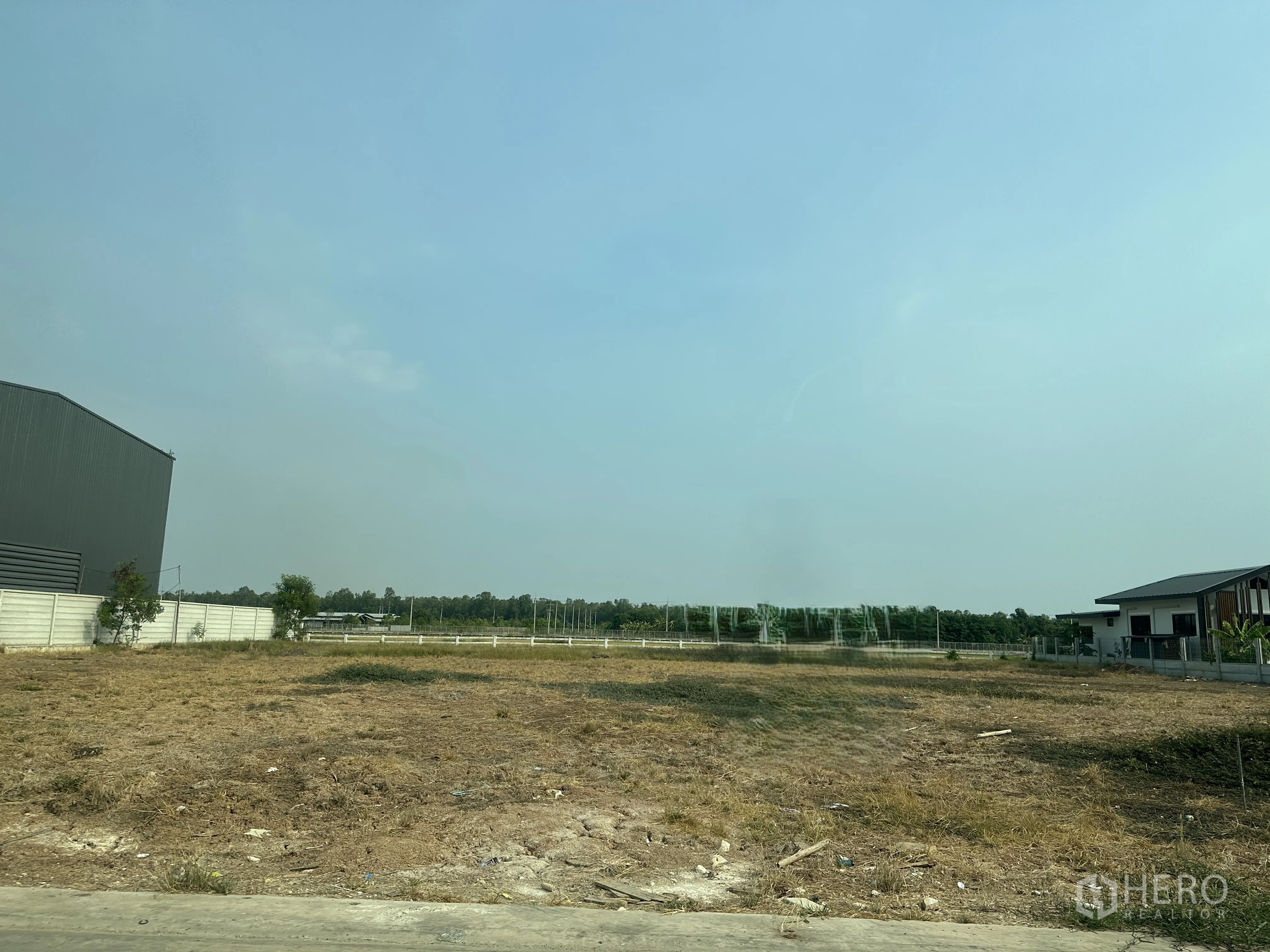 土地 for sale in Lam Luk Ka, Pathum Thani - 开阔空地，临混凝土路面，周边有围栏与邻近建筑，远处为绿地和开阔天空