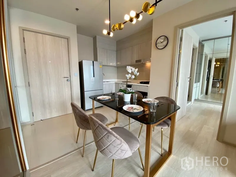 公寓 出租在Pathum Wan, Bangkok - 餐区配黑色台面餐桌与金色桌脚、绒面餐椅，旁边是白色一字型厨房与冰箱。