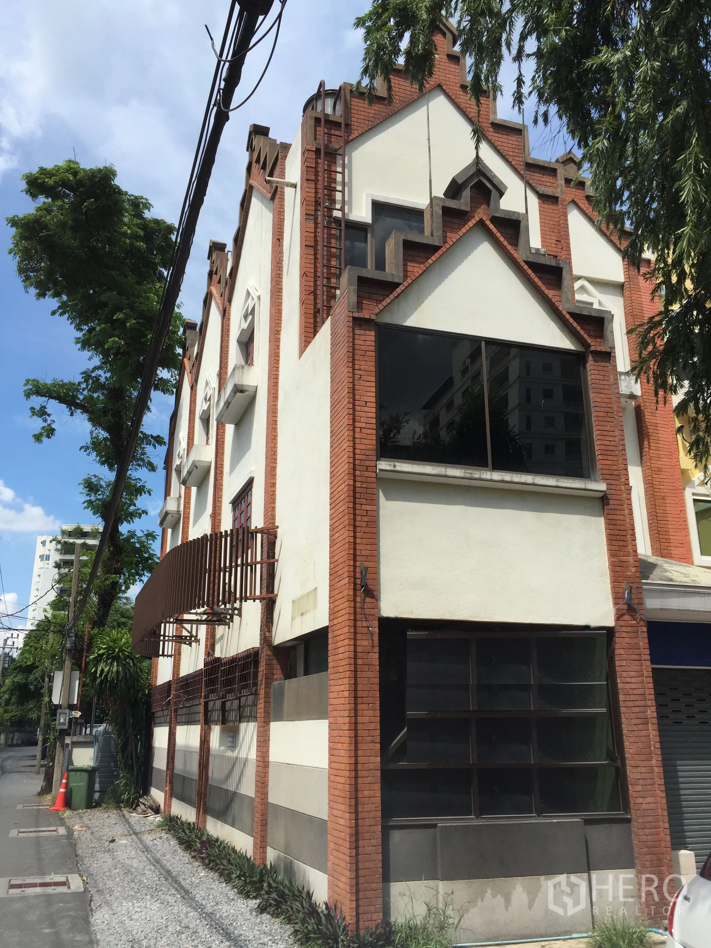 Home Office for rent in Khlong Toei, Bangkok - Corner home office with red-brick and white stepped facade, large black-framed windows, and steel awnings on a quiet tree-lined lane near BTS Thong Lo.
