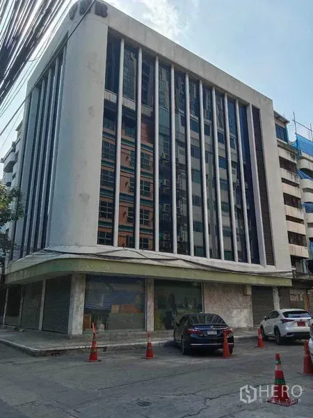 Home Office for rent in Watthana, Bangkok - Mid-rise commercial building exterior with vertical window fins and street parking in front.