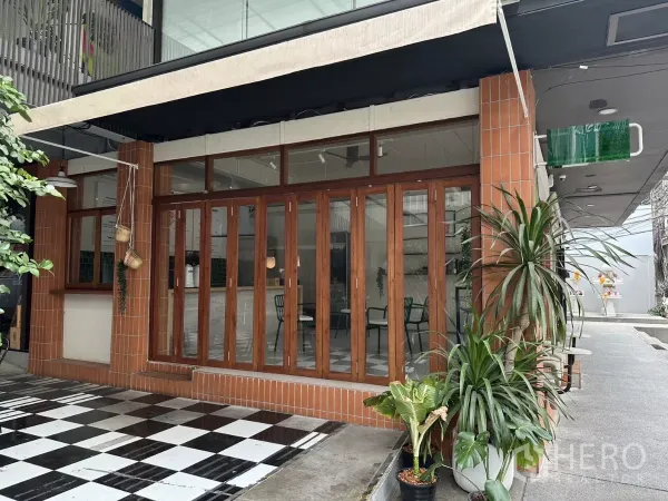 零售空间 出租在Watthana, Bangkok - 转角商铺外立面，砖柱、木框折叠玻璃门，遮阳篷与黑白棋盘格门前地面。