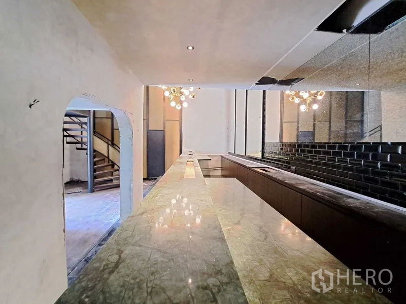 零售空间 出租在Watthana, Bangkok - 长条大理石吧台与镜面墙及吊灯，面向拱形门洞与楼梯。