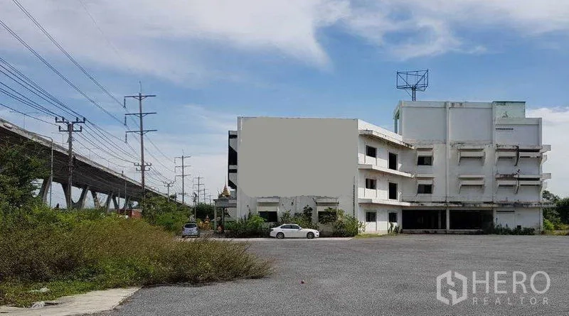 Shophouse for rent in Bang Bo, Samut Prakan - Multi-storey white commercial shophouse with expansive parking area next to an elevated roadway.