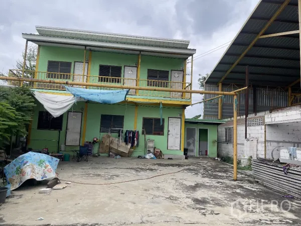 工厂 出售在Bang Bua Thong, Nonthaburi - 两层员工宿舍楼，绿色外墙，旁有硬铺院坝与棚屋。
