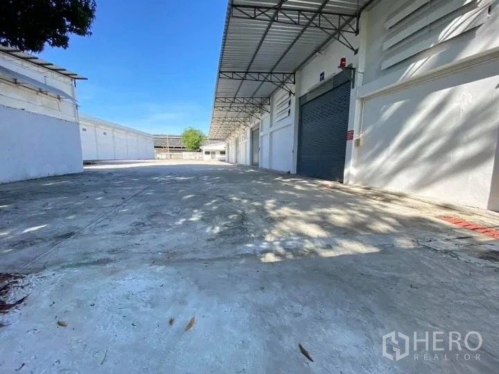 Warehouse for rent in Mueang Pathum Thani, Pathum Thani - Wide concrete yard with covered loading bays and roller shutter doors along a white warehouse.