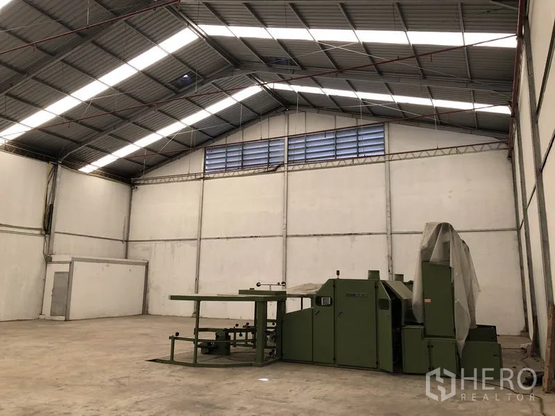 Warehouse for rent in Bang Bua Thong, Nonthaburi - High-roof warehouse corner with ridge vents and machinery on open floor.