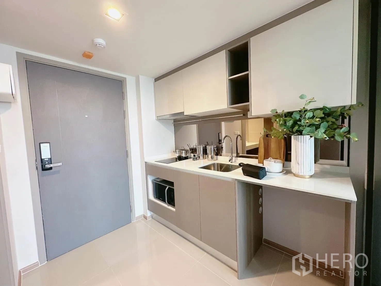 Condominium for rent in Huai Khwang, Bangkok - Compact kitchenette with white countertop, built-in cabinets, sink and induction cooktop near the entrance.