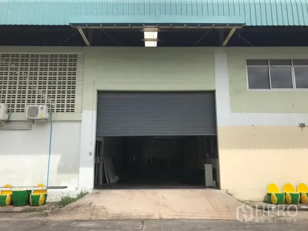 Warehouse for rent in Bang Phli, Samut Prakan - Close view of a roller shutter loading bay with concrete ramp beneath a wide canopy and adjacent windows.