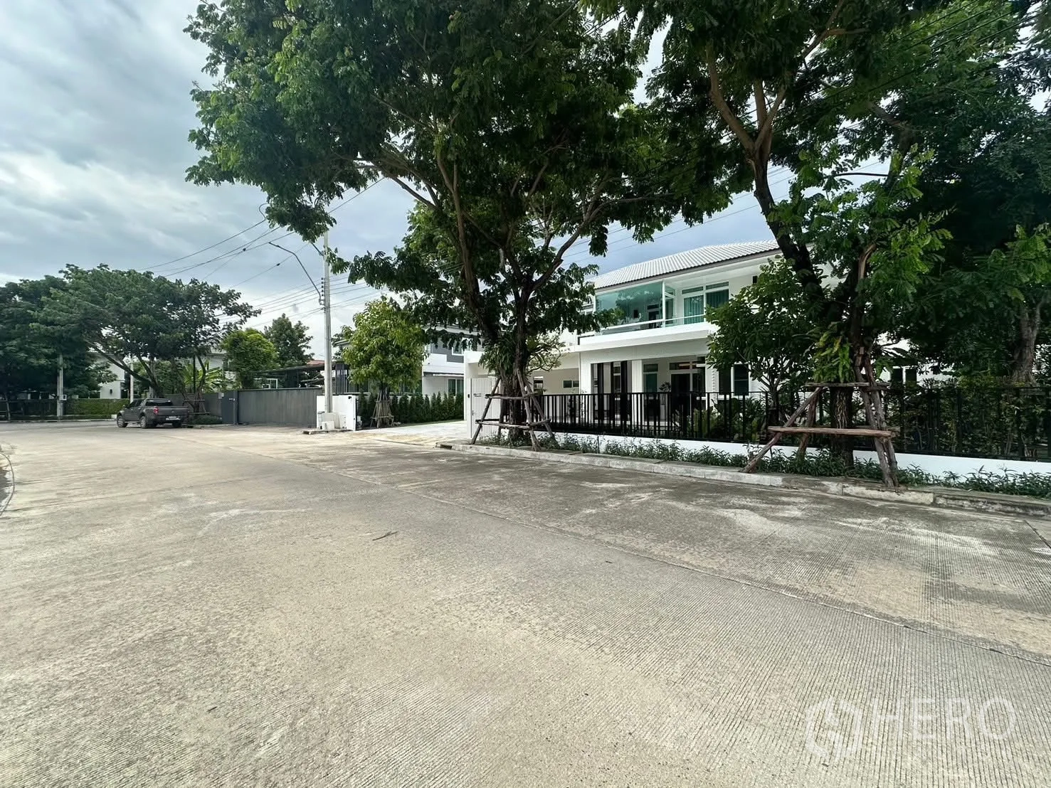 บ้านเดี่ยว for rent in Bang Phli, Samut Prakan - มุมถนนในหมู่บ้านที่ร่มรื่น เห็นรั้วสีดำและระเบียงของตัวบ้าน.