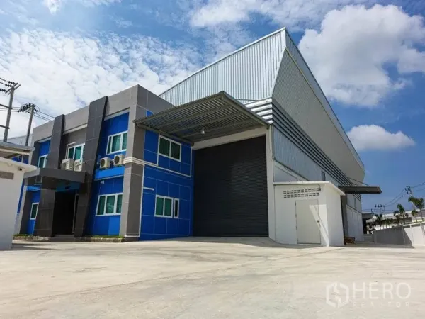 Factory for rent in Bang Bo, Samut Prakan - Modern factory exterior in Bang Bo with blue office wing, large roller shutter, covered loading bay and spacious concrete yard under a clear sky.