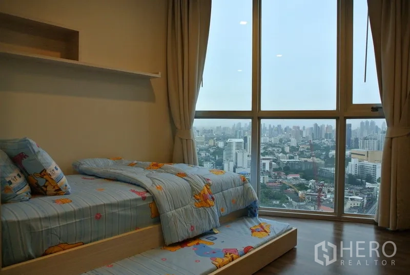 Condominium for rent or sale in Watthana, Bangkok - Cozy second bedroom with single bed and pull-out trundle, neutral curtains, wood flooring, and wide city views through floor-to-ceiling windows.