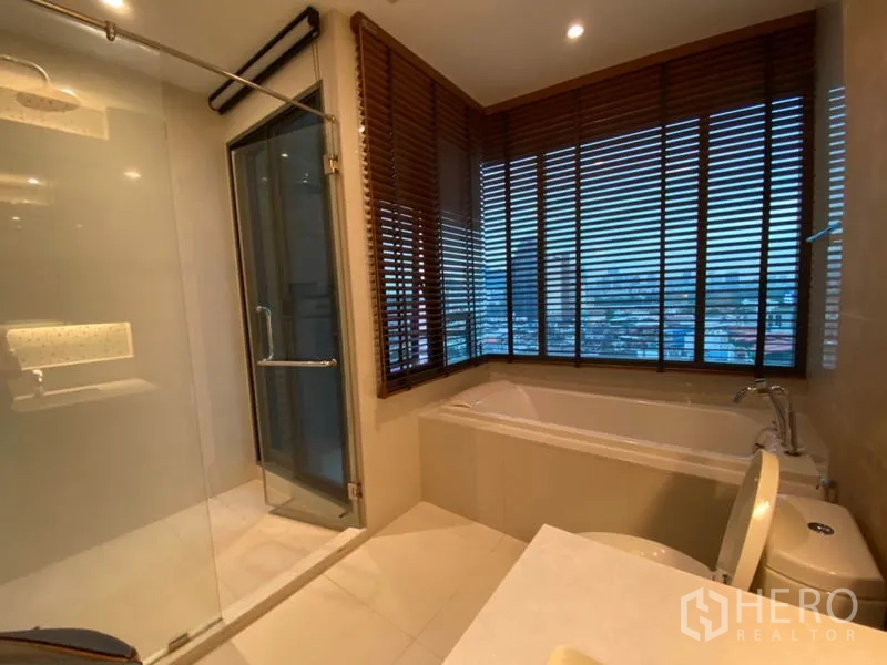 Condominium for rent in Khlong Toei, Bangkok - Bathroom with glass shower, bathtub by large windows and city skyline view.