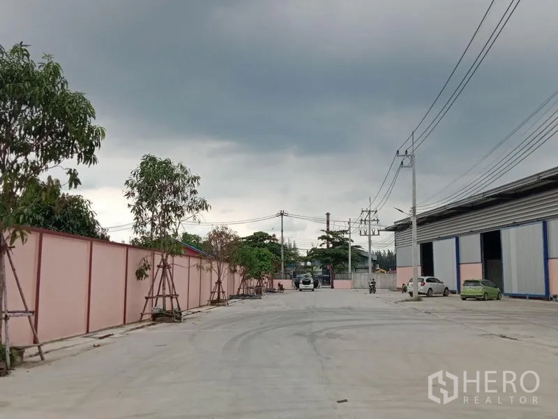 Factory for rent in Ban Phaeo, Samut Sakhon - Entrance road with landscaped trees and a secure boundary wall outside factory units.