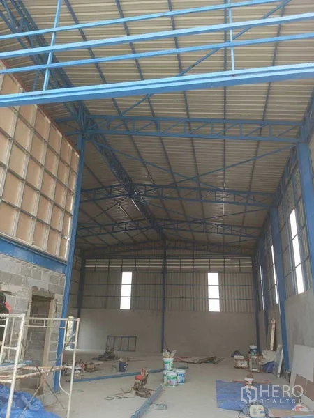 货栈 出租在Pathum Thani - 仓库内部空间开阔，屋顶高挑，可见蓝色钢桁架与地面施工材料。
