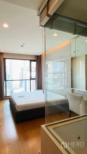 Condominium for rent or sale in Bang Rak, Bangkok - Bedroom with floor-to-ceiling windows and skyline view, glass wall to the bathroom and a small desk.