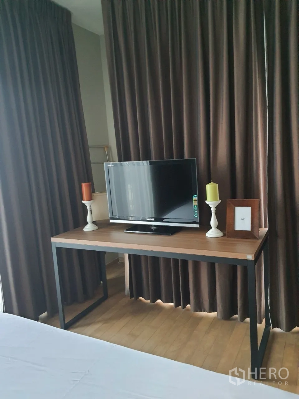 Condominium for rent or sale in Watthana, Bangkok - Bedroom corner with TV on a wooden desk, floor-to-ceiling dark curtains and wood flooring.