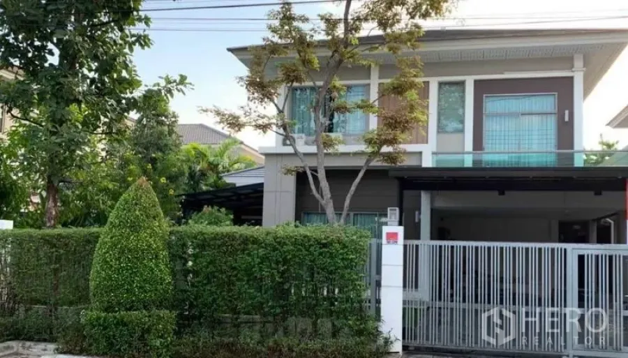 独立式住宅 出售在Mueang Pathum Thani, Pathum Thani - 房屋正立面，两层独栋别墅配绿篱围栏、滑动大门与有顶车棚。