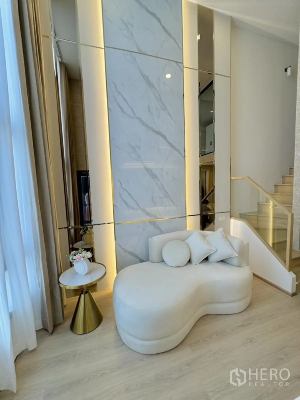 公寓 for rent in Khlong Toei, Bangkok - 客厅设奶油色躺榻、背光大理石特色墙、大窗与浅色木地板，并有玻璃扶手楼梯通向上层。