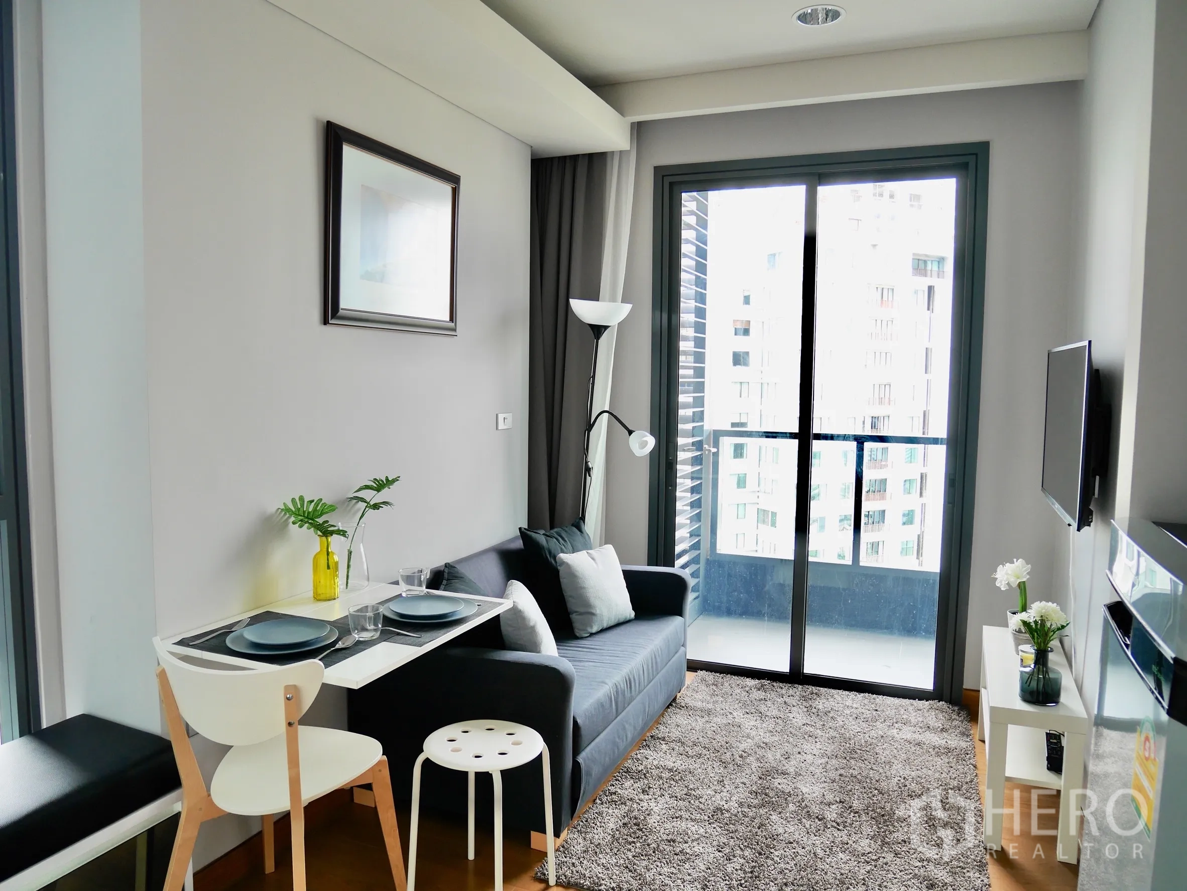 Condominium for rent in Khlong Toei, Bangkok - Bright living room with gray sofa, wall-mounted TV, rug, and sliding doors opening to a balcony with city view.